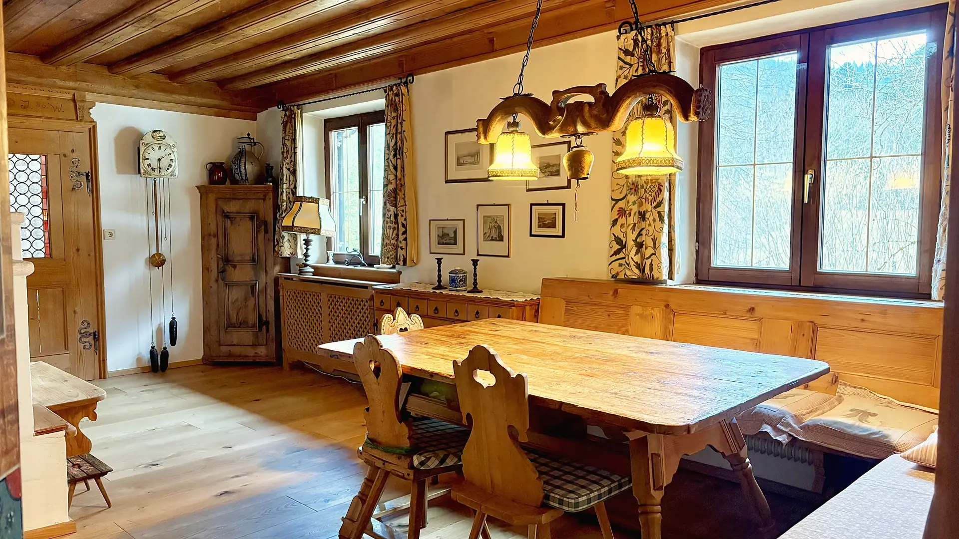 Langer Holztisch mit geschnitzten Bauernstühlen unter zwei Schmiedeleuchten, ringsum Holzvertäfelung und eine alte Standuhr.