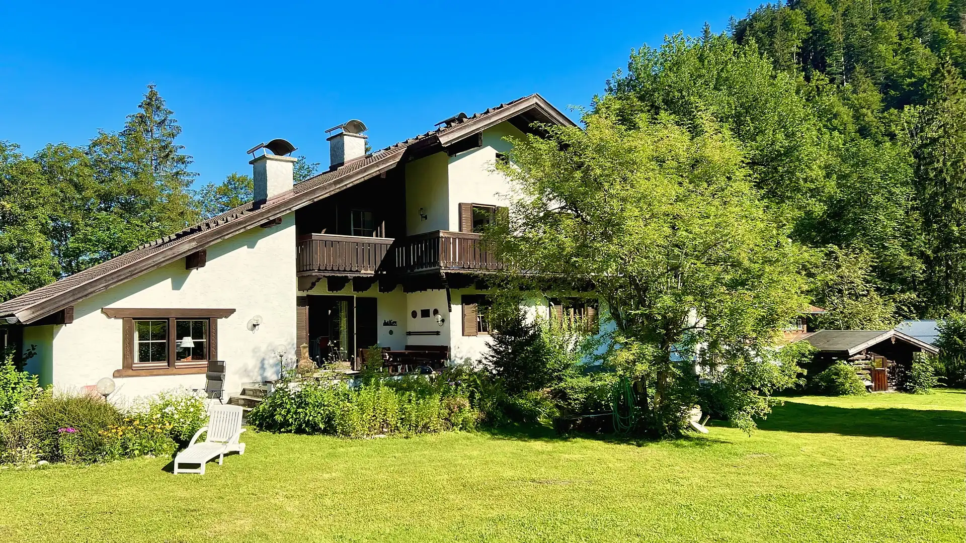 Das Ferienhaus von der Gartenseite an einem Sommertag: heller Putz, Holzbalkon, dunkles Satteldach, davor eine große Wiese, dahinter ein bewaldeter Hang unter blauem Himmel.
