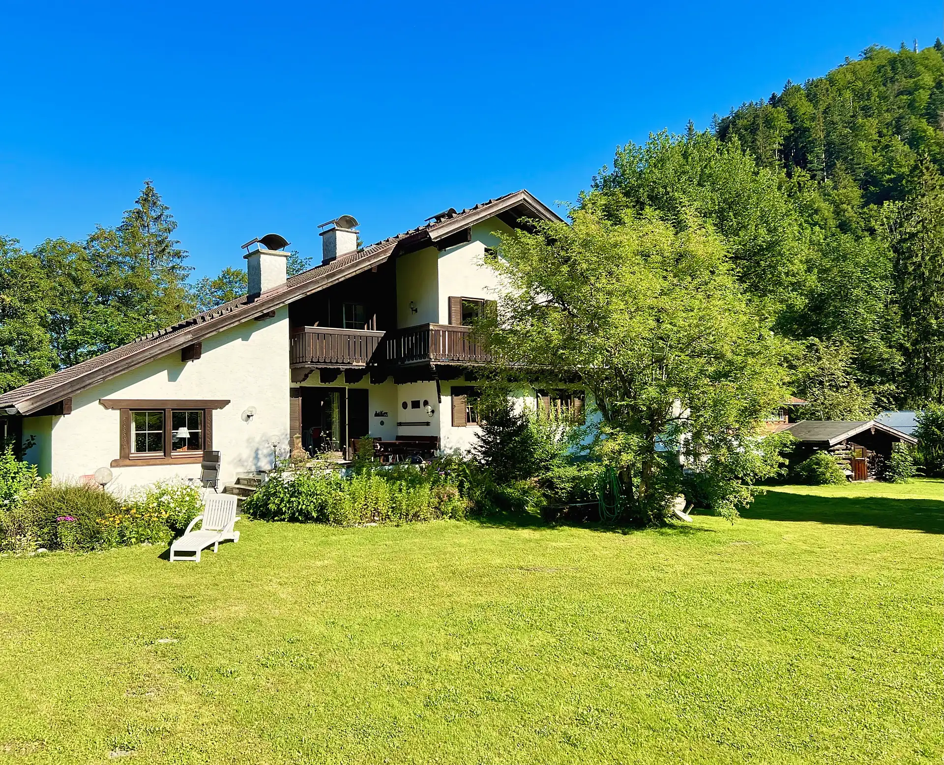 Das Ferienhaus von der Gartenseite an einem Sommertag: heller Putz, Holzbalkon, dunkles Satteldach, davor eine große Wiese, dahinter ein bewaldeter Hang unter blauem Himmel.