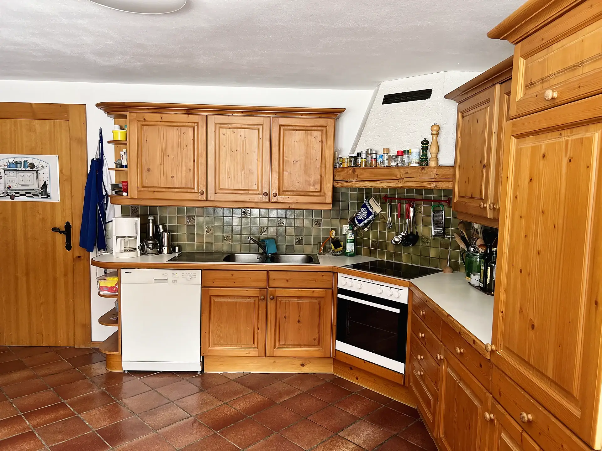 Küchenzeile über Eck mit grünen Wandfliesen, Ceranfeld, Backofen, Geschirrspüler und Spüle.