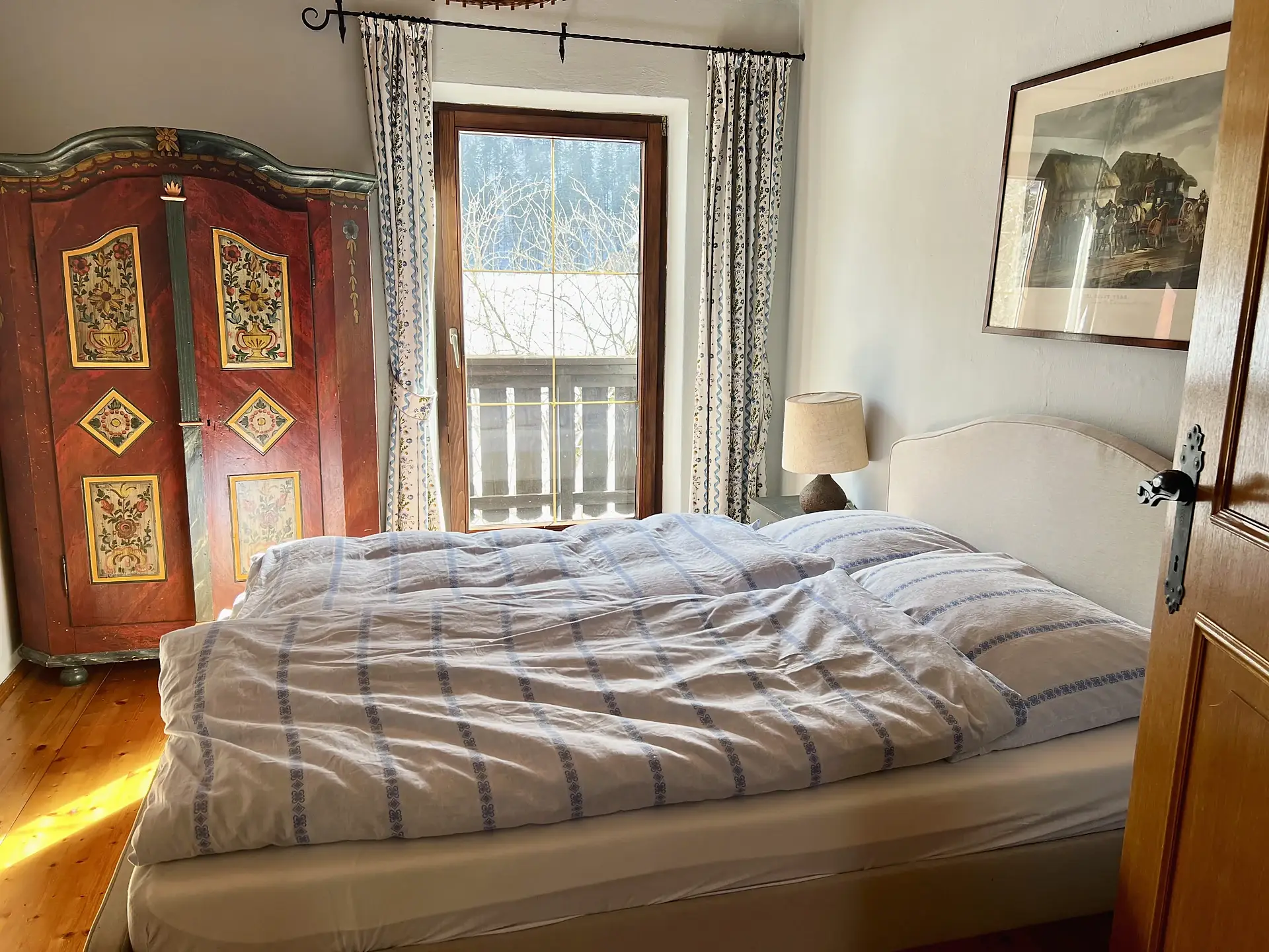 Schlafzimmer mit rot bemaltem antiken Bauernschrank, Doppelbett und Tür zum Balkon.