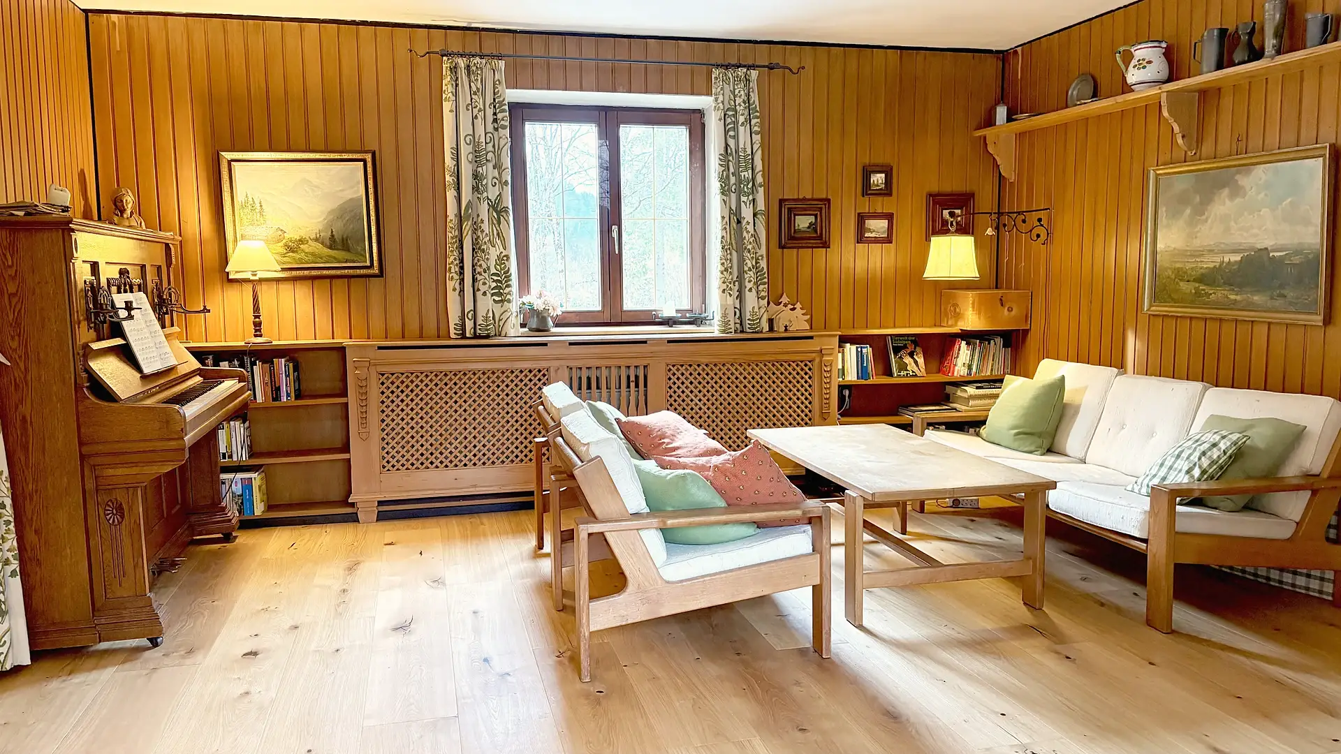 Gleicher Raum, Blick auf die Sitzgruppe vor dem Fenster und die Bilder an der Holzwand.