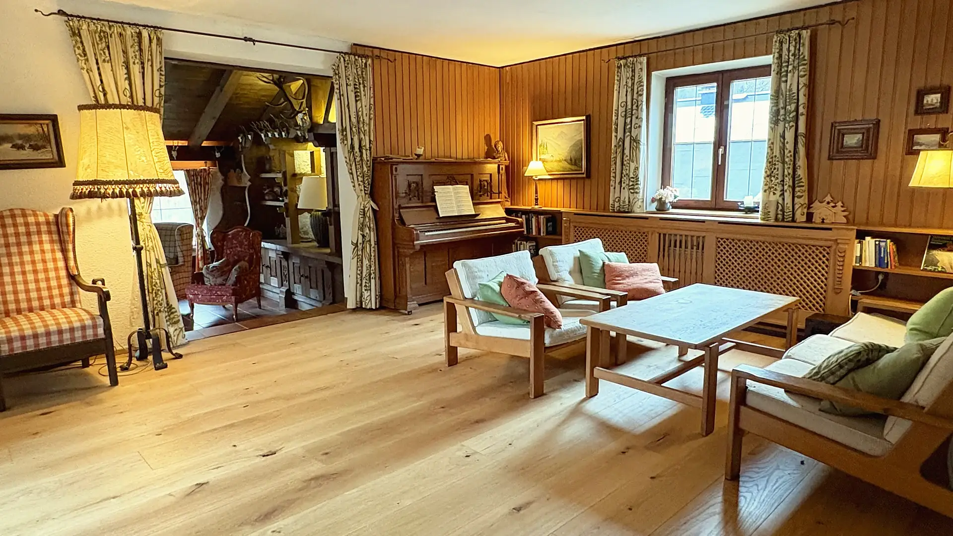 Holzvertäfelter Wohnraum mit Klavier, Bücherregal und hellen Polstermöbeln um einen niedrigen Holztisch.