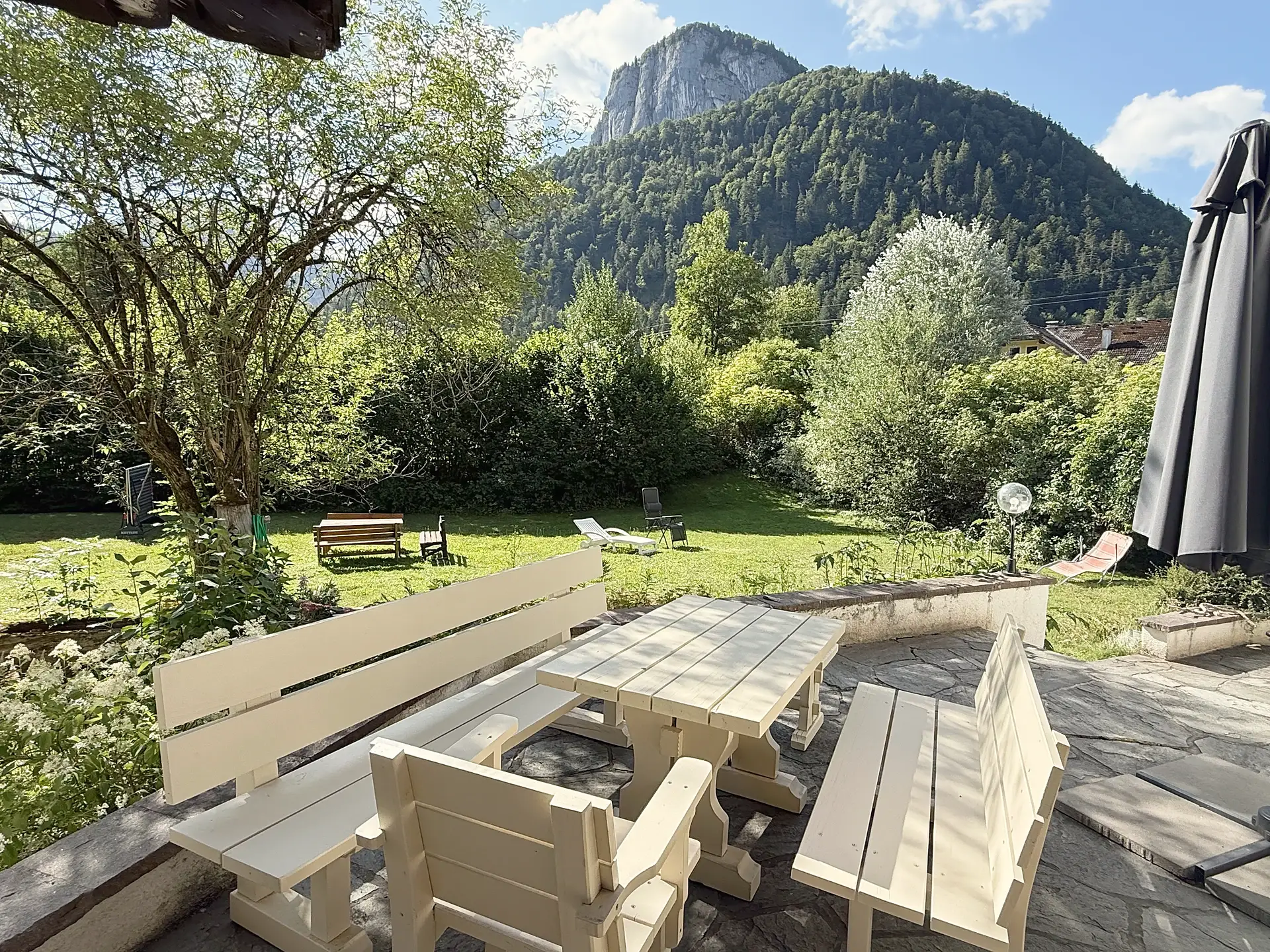 Terrasse mit hellen Holzbänken und Tisch, dahinter der Garten und eine bewaldete Bergflanke mit heller Felswand.