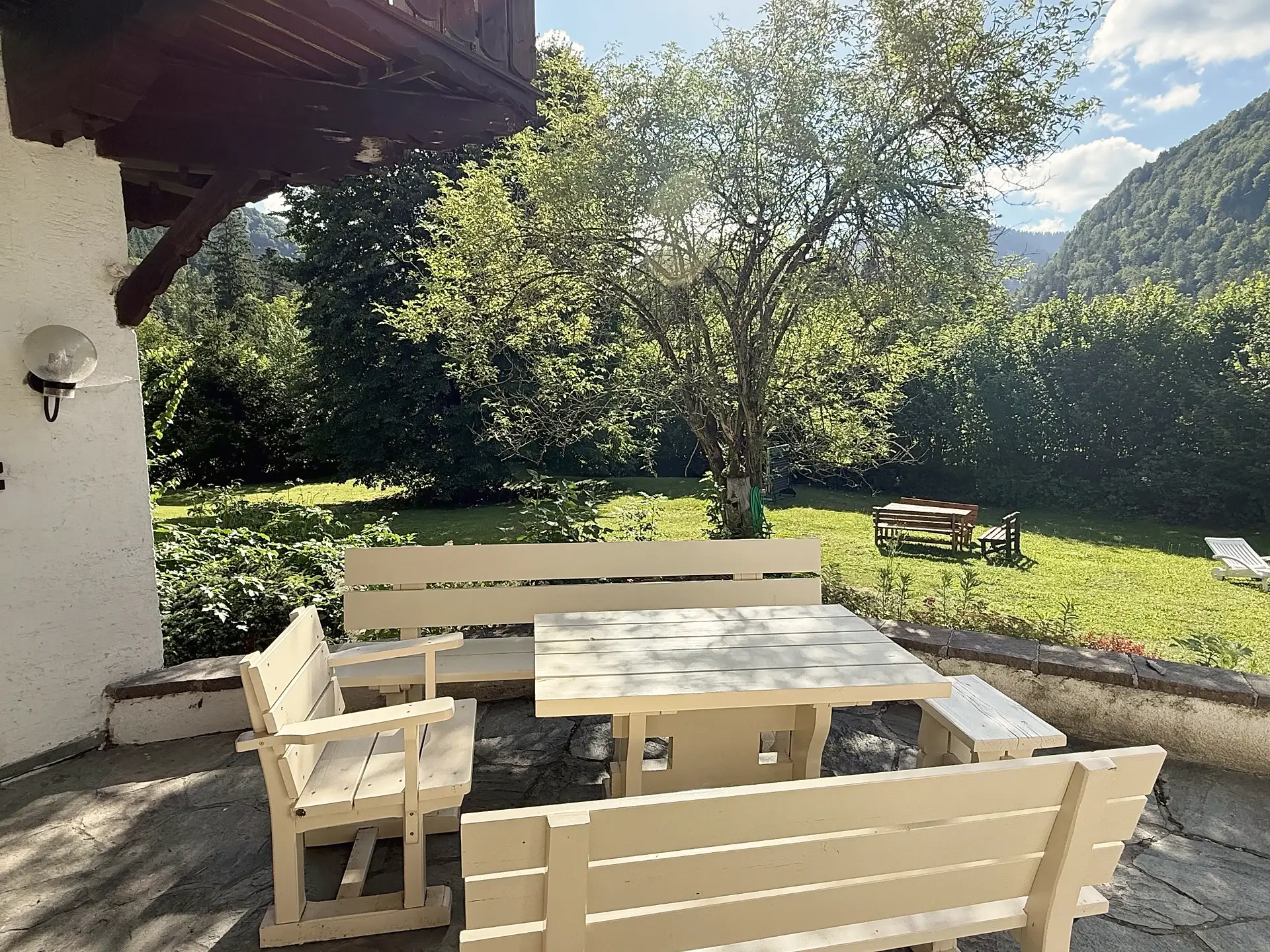 Überdachte Terrasse mit hellen Holzbänken und Tisch, dahinter der Garten mit altem Obstbaum und bewaldeten Hängen.